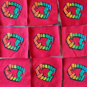 May include: Nine square red fabric patches arranged in a grid, each featuring a central graphic of a set of vampire fangs. The fangs are outlined in black and filled with a gradient of orange, yellow, green, and blue, with red glitter accents.