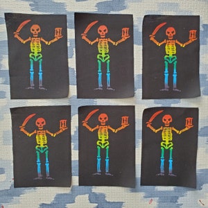 May include: Six black fabric patches with a colourful skeleton design. The skeletons are in a rainbow gradient, holding a sword and an hourglass. The patches are arranged on a light blue and white patterned surface.