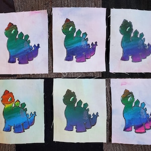 Könnte beinhalten: Sechs bunte Stoffquadrate mit einem Regenbogen-Dinosaurier-Design. Jedes Quadrat hat ein anderes Farbschema, wobei der Körper und der Schwanz des Dinosauriers in einen Farbverlauf übergehen.