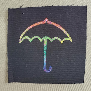 Pode incluir: Um patch quadrado de tecido preto com um design de guarda-chuva arco-íris brilhante. O contorno do guarda-chuva é um gradiente de cores, criando uma imagem vibrante. O patch é provavelmente para costurar ou passar a ferro em roupas ou acessórios.