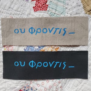 May include: Two rectangular fabric patches. The top patch is beige with blue Greek text. The bottom patch is black with the same blue Greek text. The patches are on a patchwork quilt.
