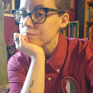 May include: A person wearing a red and white striped button-down shirt with a penguin patch on the left side. The person has a tattoo on their left forearm that reads "Truth is Out There".