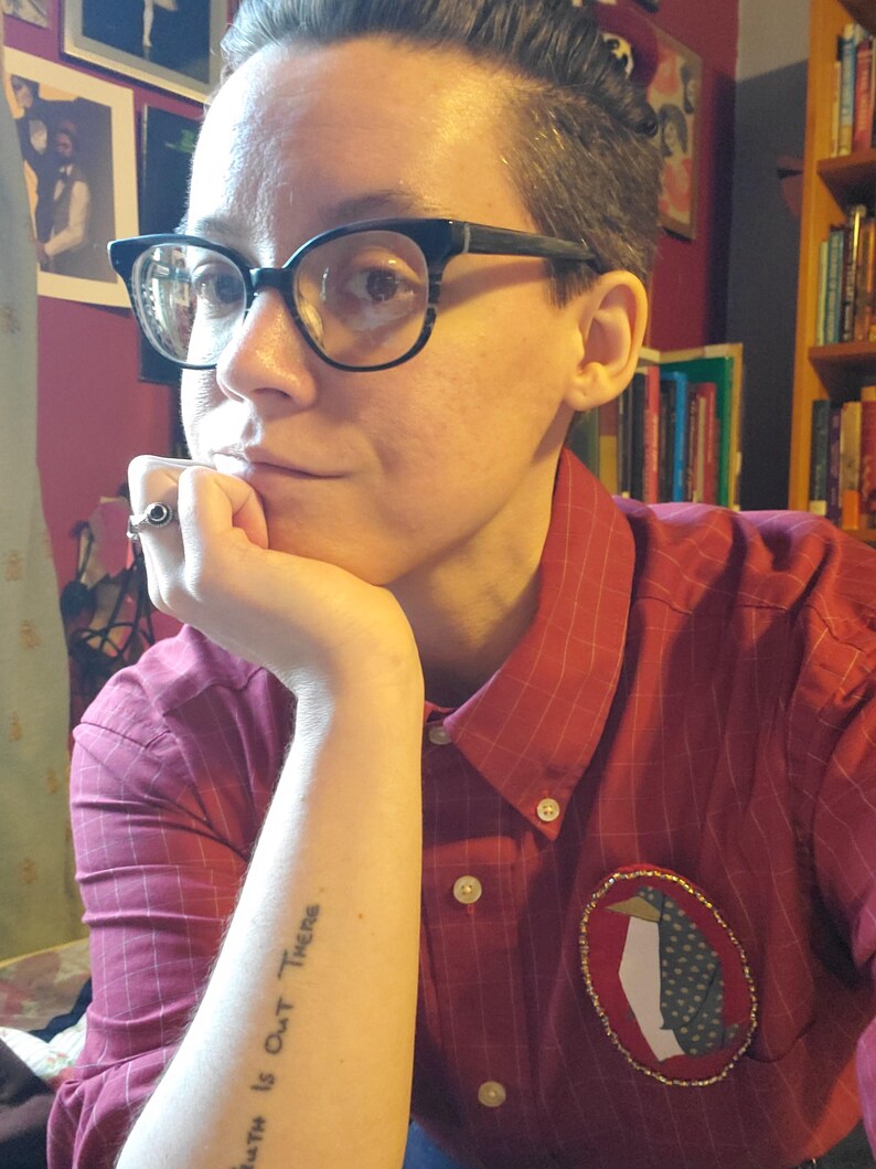 May include: A person wearing a red and white striped button-down shirt with a penguin patch on the left side. The person has a tattoo on their left forearm that reads "Truth is Out There".