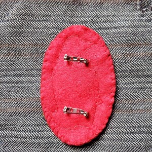 May include: The back of a red felt pin with two silver safety pins attached.