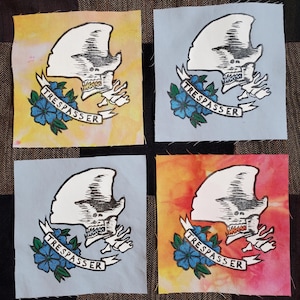 May include: Four fabric squares featuring a white alien skull graphic and the word "TRESPASSER" in a banner. The squares are in four different colours: yellow, blue, grey, and red.