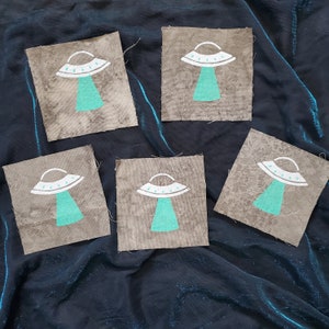 Op de afbeelding: Vijf grijze stoffen patches met een wit en turquoise UFO-ontwerp. Elke patch heeft een eenvoudig UFO met een afgeronde bovenkant en een turquoise lichtstraal.