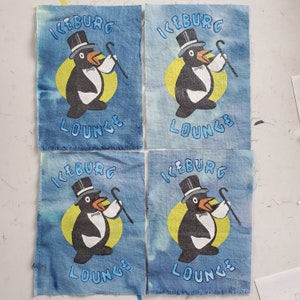 May include: Four blue fabric squares with a white penguin design. Each square has the text "Iceburg Lounge" printed on it.