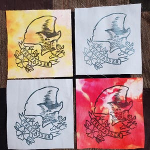 May include: Four fabric squares featuring a black and white print of a skull wearing a hat and a banner that reads "TRESPASSER". The squares are in different colours: yellow, white, red, and white.