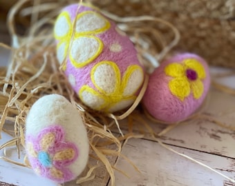 May include: Three felt Easter eggs decorated with pink and yellow flowers. The eggs are sitting on a wooden surface with straw.