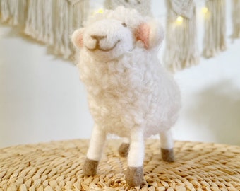 Pode incluir: Um brinquedo de ovelha branco e felpudo com orelhas cor-de-rosa e pés castanhos. A ovelha está em pé sobre uma superfície de vime trançada.
