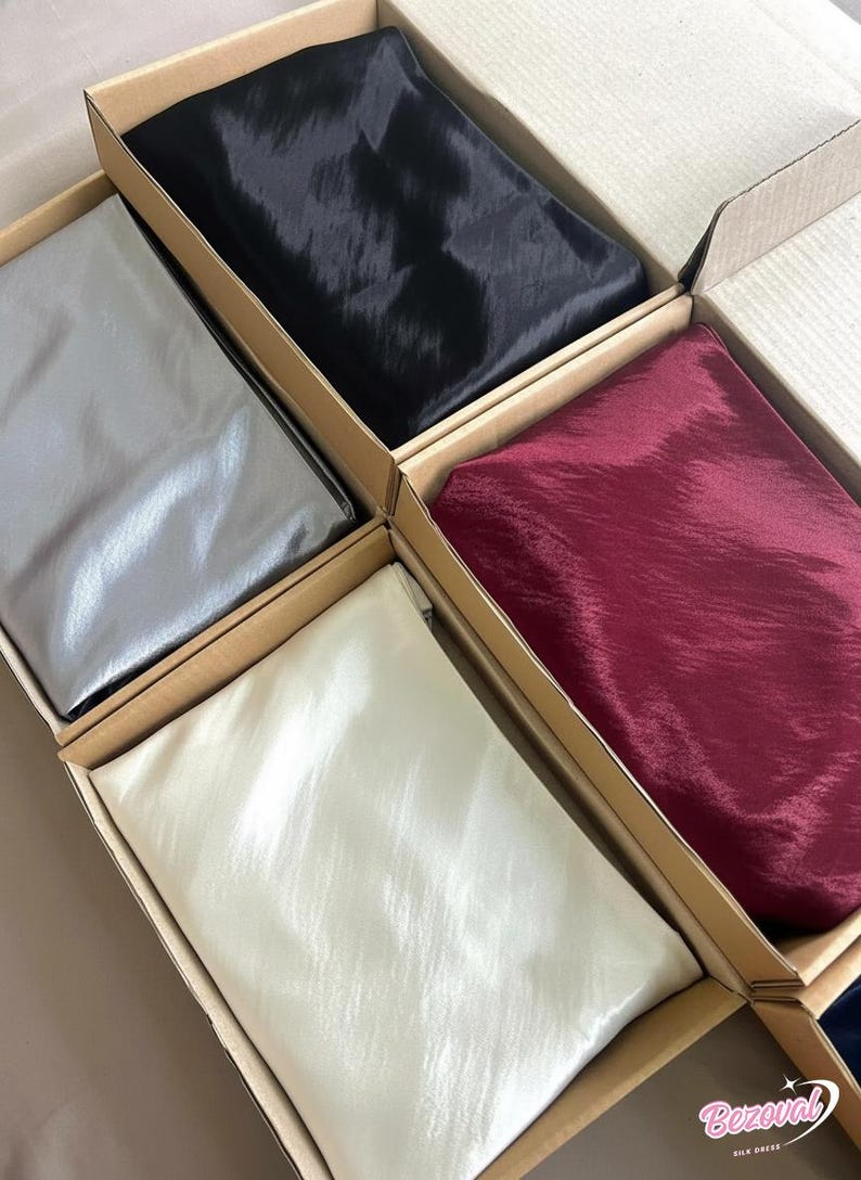 May include: An assortment of silk scarves in cardboard boxes. The scarves are in various colors, including black, silver, burgundy, and cream. The silk has a subtle sheen, and the boxes are arranged in a grid pattern.