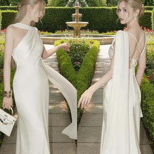 Puede incluir: Dos mujeres con elegantes vestidos blancos posan al aire libre. Los vestidos presentan escotes asim&eacute;tricos &uacute;nicos y detalles de tela fluida. Uno tiene un hombro drapeado, el otro una espalda cruzada. El fondo incluye una fuente y vegetaci&oacute;n cuidada.