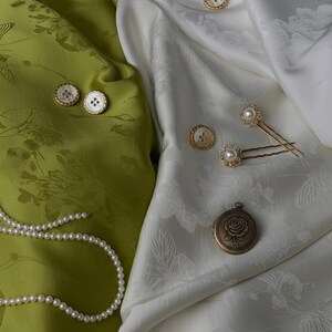May include: An assortment of vintage-style jewelry and accessories, including pearl necklaces, buttons, hairpins, and a locket. The items are arranged on green and white floral patterned fabric. The locket features a rose design.