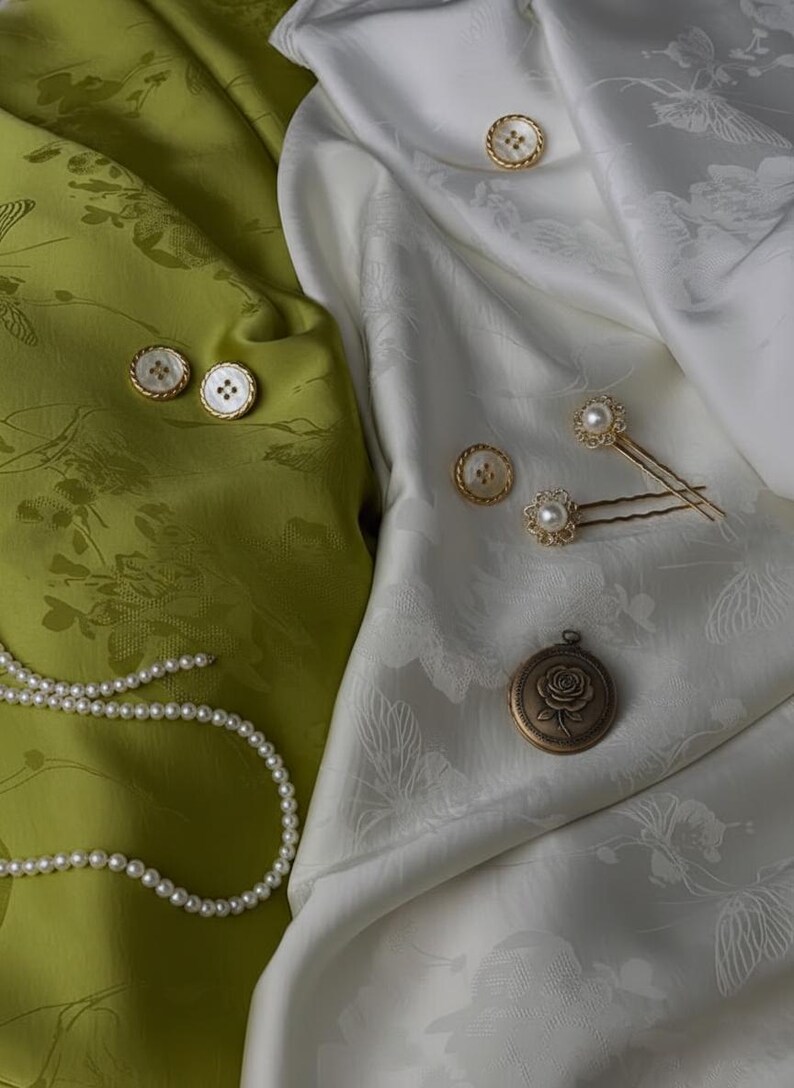 May include: An assortment of vintage-style jewelry and accessories, including pearl necklaces, buttons, hairpins, and a locket. The items are arranged on green and white floral patterned fabric. The locket features a rose design.