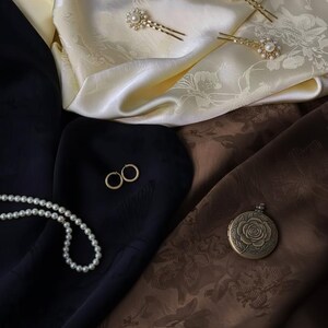 May include: An assortment of jewelry and accessories displayed on a cream and brown patterned fabric. The items include gold hairpins with pearl accents, gold hoop earrings, a pearl necklace, and a round locket with a rose design.