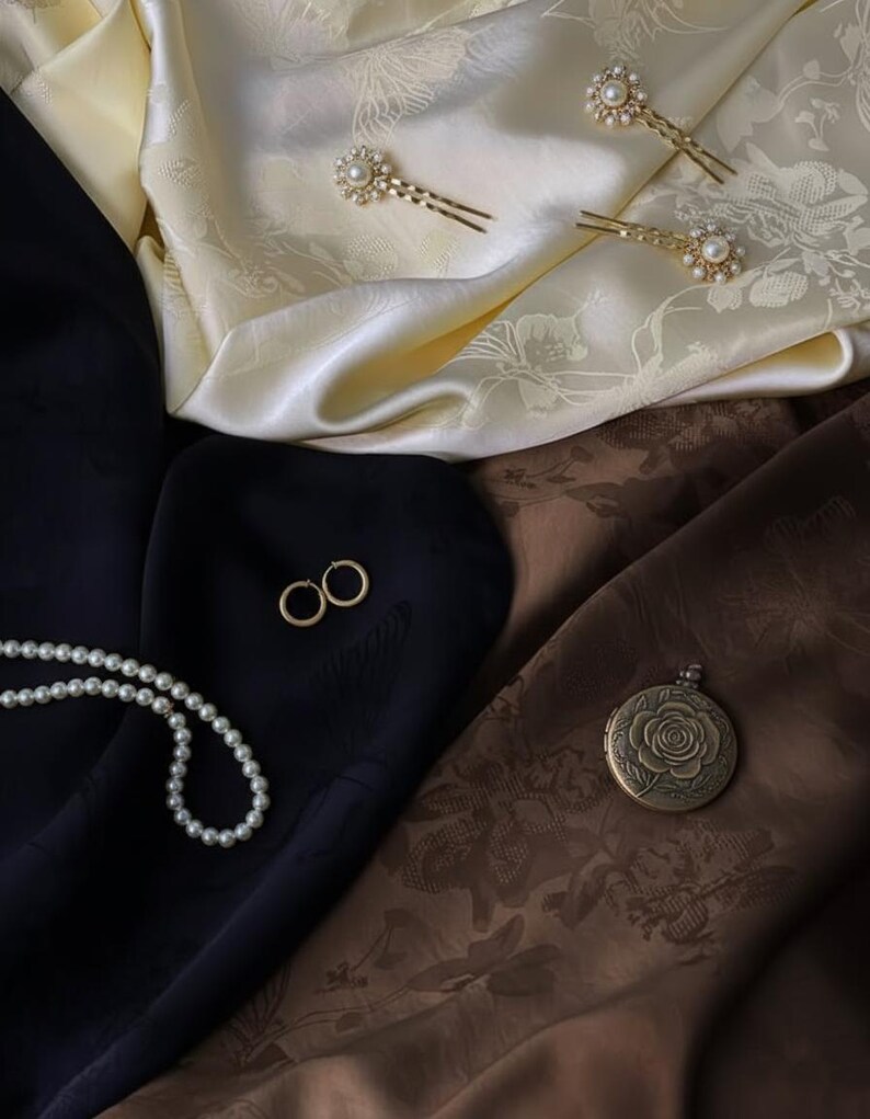 May include: An assortment of jewelry and accessories displayed on a cream and brown patterned fabric. The items include gold hairpins with pearl accents, gold hoop earrings, a pearl necklace, and a round locket with a rose design.