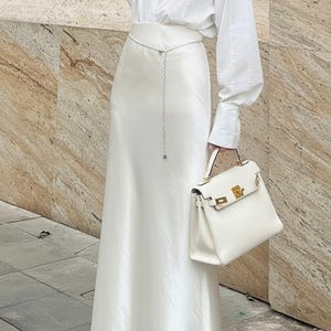 May include: An all-white ensemble featuring a long satin skirt, a button-down shirt, and a structured handbag. A silver chain belt adds a subtle accent. The outfit is completed with silver heeled sandals. The image is taken outdoors.