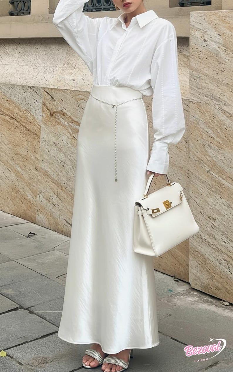 May include: An all-white ensemble featuring a long satin skirt, a button-down shirt, and a structured handbag. A silver chain belt adds a subtle accent. The outfit is completed with silver heeled sandals. The image is taken outdoors.