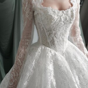 Puede incluir: Vestido de novia blanco con intrincados detalles de encaje. El vestido presenta un escote cuadrado, mangas largas y una falda amplia. El corpi&ntilde;o es entallado y el encaje se extiende sobre los hombros. Vestido de novia Bezoval.