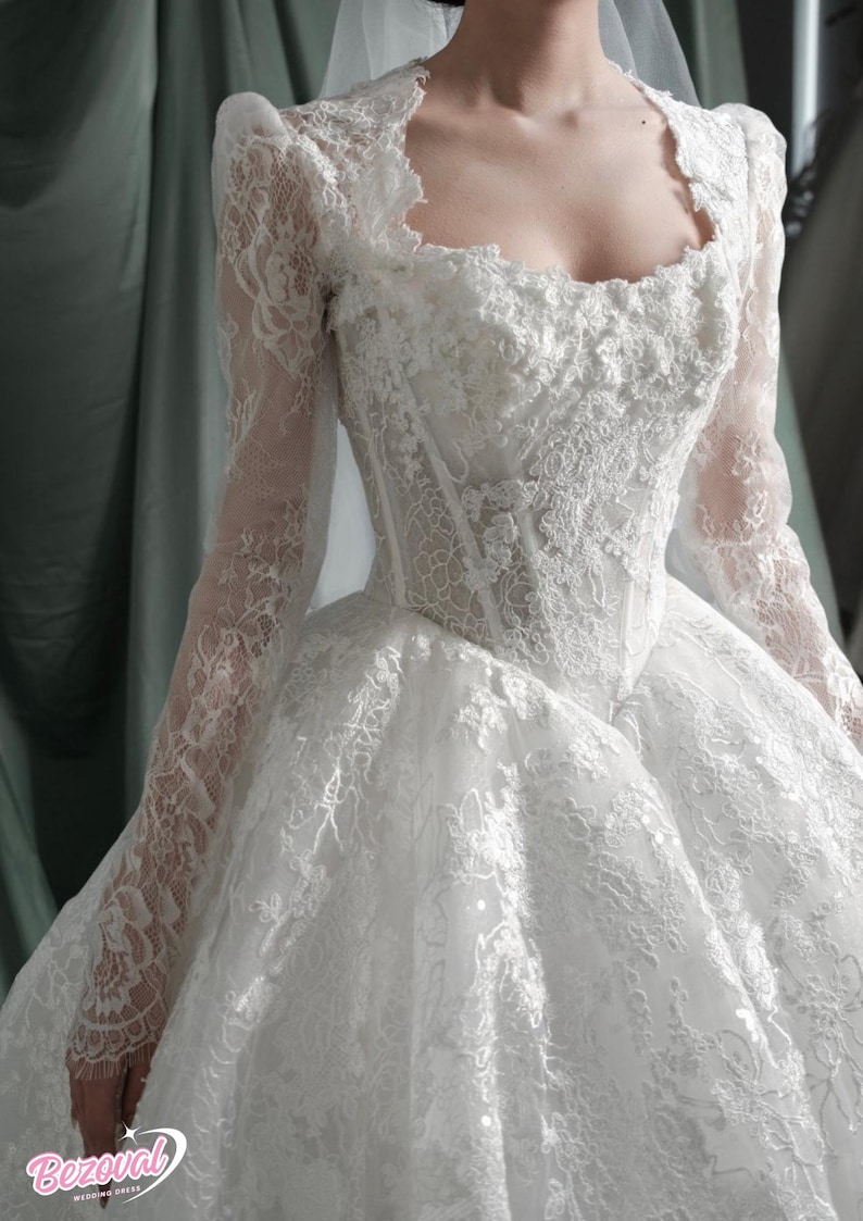 Puede incluir: Vestido de novia blanco con intrincados detalles de encaje. El vestido presenta un escote cuadrado, mangas largas y una falda amplia. El corpi&ntilde;o es entallado y el encaje se extiende sobre los hombros. Vestido de novia Bezoval.