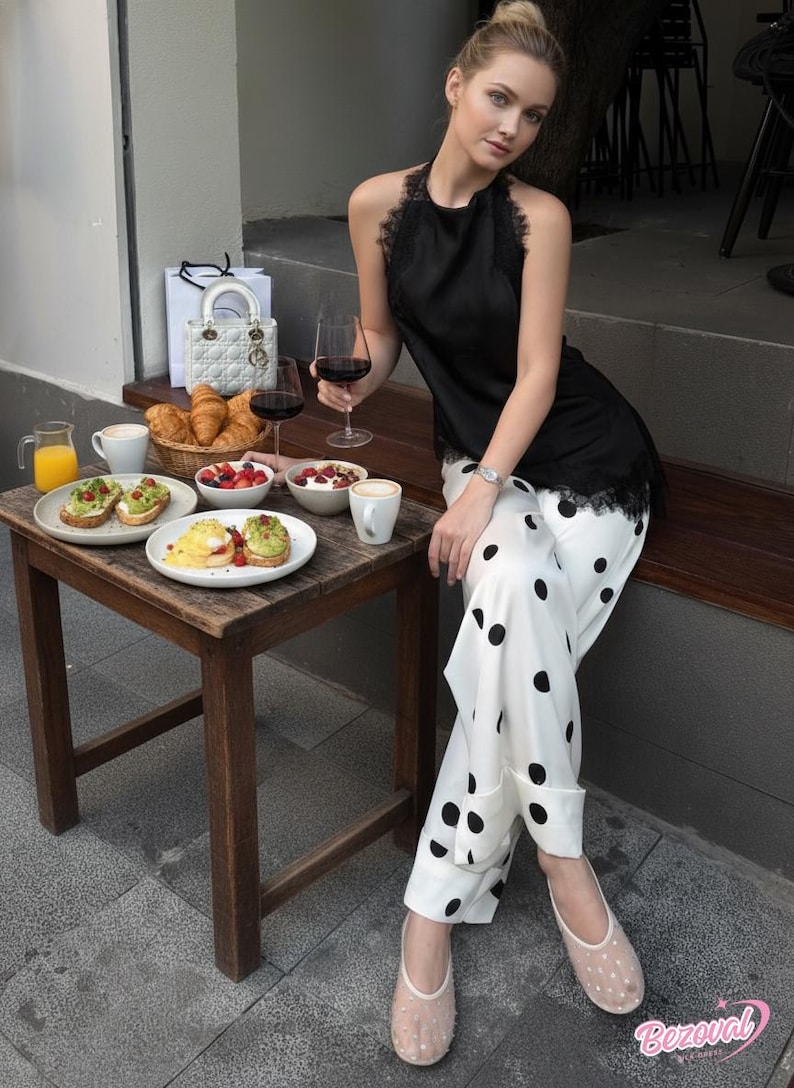 May include: A woman wearing a black halter top with lace trim and white polka dot pants. A small wooden table holds a basket of croissants, plates of food, and glasses of red wine. A white handbag sits nearby.