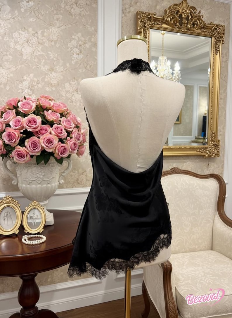 May include: A black halter top with lace trim is displayed on a mannequin. The top has a low back and a delicate lace detail around the neckline. The mannequin is in a room with a floral wallpaper and a gold-framed mirror.