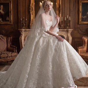 Puede incluir: Vestido de novia blanco sin tirantes con intrincados detalles de encaje y una falda amplia. La novia lleva un velo transparente y est&aacute; sentada en una habitaci&oacute;n con una decoraci&oacute;n ornamentada.