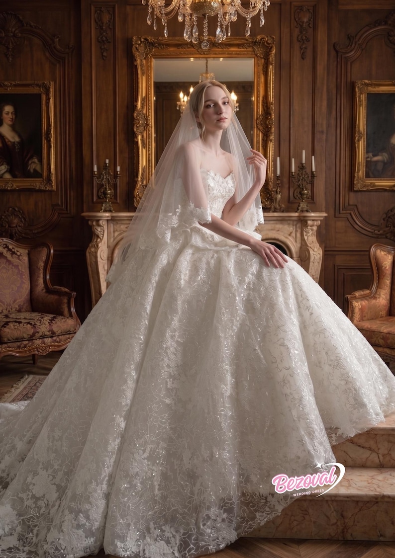 Puede incluir: Vestido de novia blanco sin tirantes con intrincados detalles de encaje y una falda amplia. La novia lleva un velo transparente y est&aacute; sentada en una habitaci&oacute;n con una decoraci&oacute;n ornamentada.
