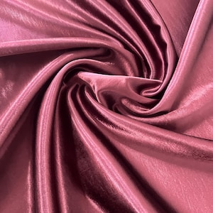 May include: Close-up of a swirl of mauve-colored satin fabric. The fabric has a smooth, shiny texture with visible folds and highlights. The material appears to be silk or a silk blend, suitable for apparel or decorative projects.