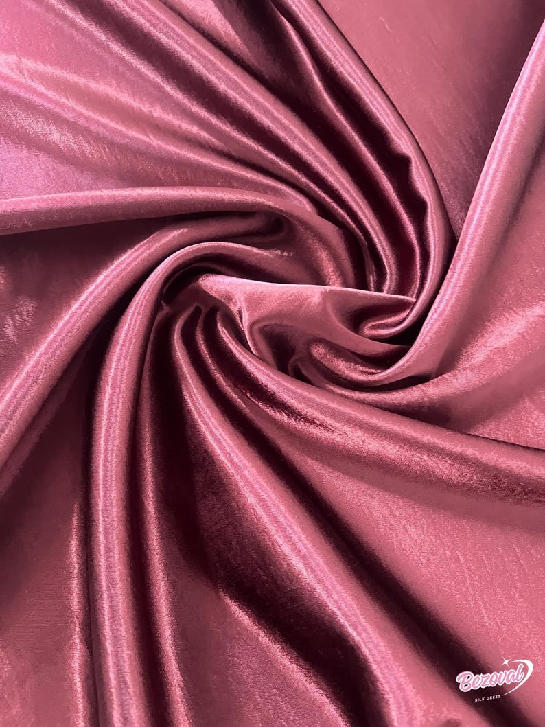 May include: Close-up of a swirl of mauve-colored satin fabric. The fabric has a smooth, shiny texture with visible folds and highlights. The material appears to be silk or a silk blend, suitable for apparel or decorative projects.