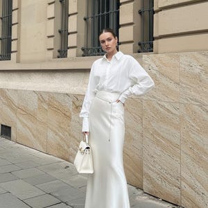 May include: A woman wearing a white button-down shirt and a long, white satin skirt. She is holding a white handbag and wearing white sandals. The background features a building with windows and stone facade.