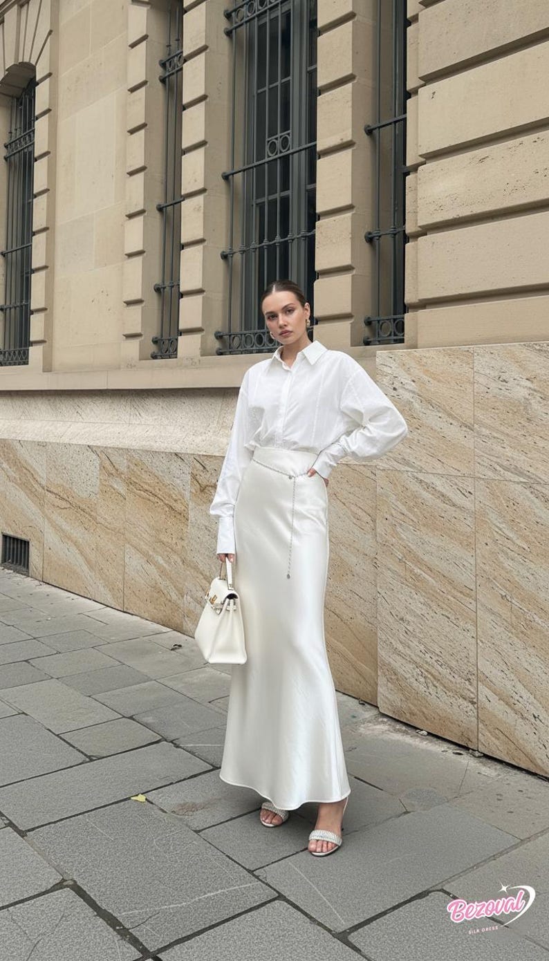 May include: A woman wearing a white button-down shirt and a long, white satin skirt. She is holding a white handbag and wearing white sandals. The background features a building with windows and stone facade.