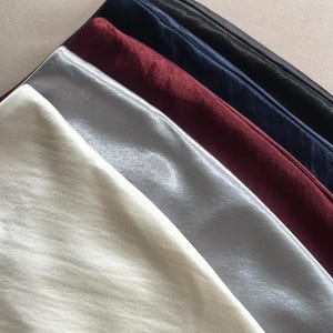 May include: An assortment of satin fabric in various colors, including ivory, silver, burgundy, navy, and black. The fabric is folded and arranged diagonally, showcasing the smooth texture and sheen of the material. The image is well-lit, highlighting the different shades.