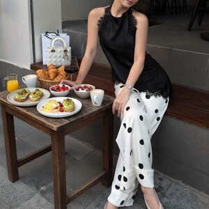 May include: A woman wearing a black halter top with lace trim and white polka dot pants. A small wooden table holds breakfast items, including avocado toast, fruit, and coffee. A white handbag and a glass of red wine are also present.