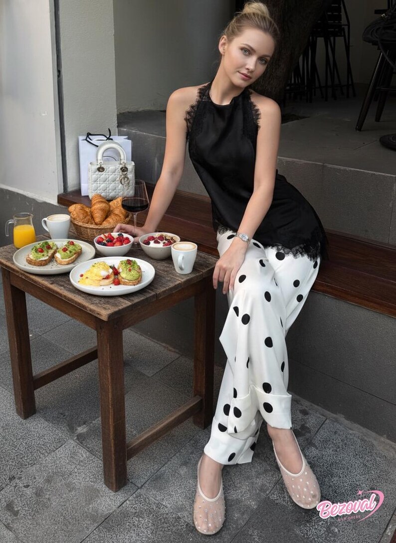 May include: A woman wearing a black halter top with lace trim and white polka dot pants. A small wooden table holds breakfast items, including avocado toast, fruit, and coffee. A white handbag and a glass of red wine are also present.