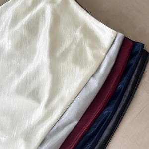 May include: A collection of fabric swatches in various colors, including off-white, silver, burgundy, navy blue, and black. The fabric has a shiny, slightly textured appearance, suggesting a satin or similar material. The swatches are arranged in a diagonal line.