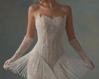 May include: A strapless, ivory-colored mini dress featuring intricate lace embroidery, beadwork, and a corset-style bodice. The garment is adorned with long, dangling beaded fringe along the hemline and includes matching lace opera-length gloves.