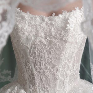 Puede incluir: Vestido de novia blanco con intrincados detalles de encaje. El corpi&ntilde;o presenta patrones florales y un escote coraz&oacute;n. Un velo transparente oscurece parcialmente el vestido. El vestido est&aacute; adornado con lentejuelas y peque&ntilde;as cuentas.