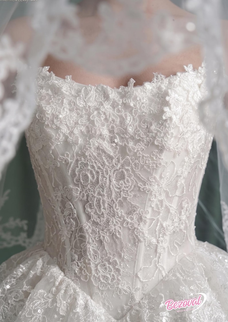 Puede incluir: Vestido de novia blanco con intrincados detalles de encaje. El corpi&ntilde;o presenta patrones florales y un escote coraz&oacute;n. Un velo transparente oscurece parcialmente el vestido. El vestido est&aacute; adornado con lentejuelas y peque&ntilde;as cuentas.