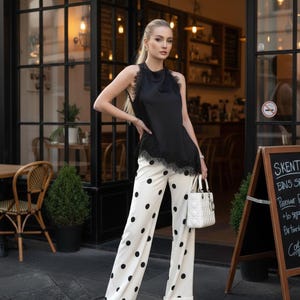 May include: A woman's outfit featuring a black sleeveless top with lace trim and white polka dot pants. The pants have a wide leg design. A white handbag and beige shoes complete the look. The background includes a cafe setting.