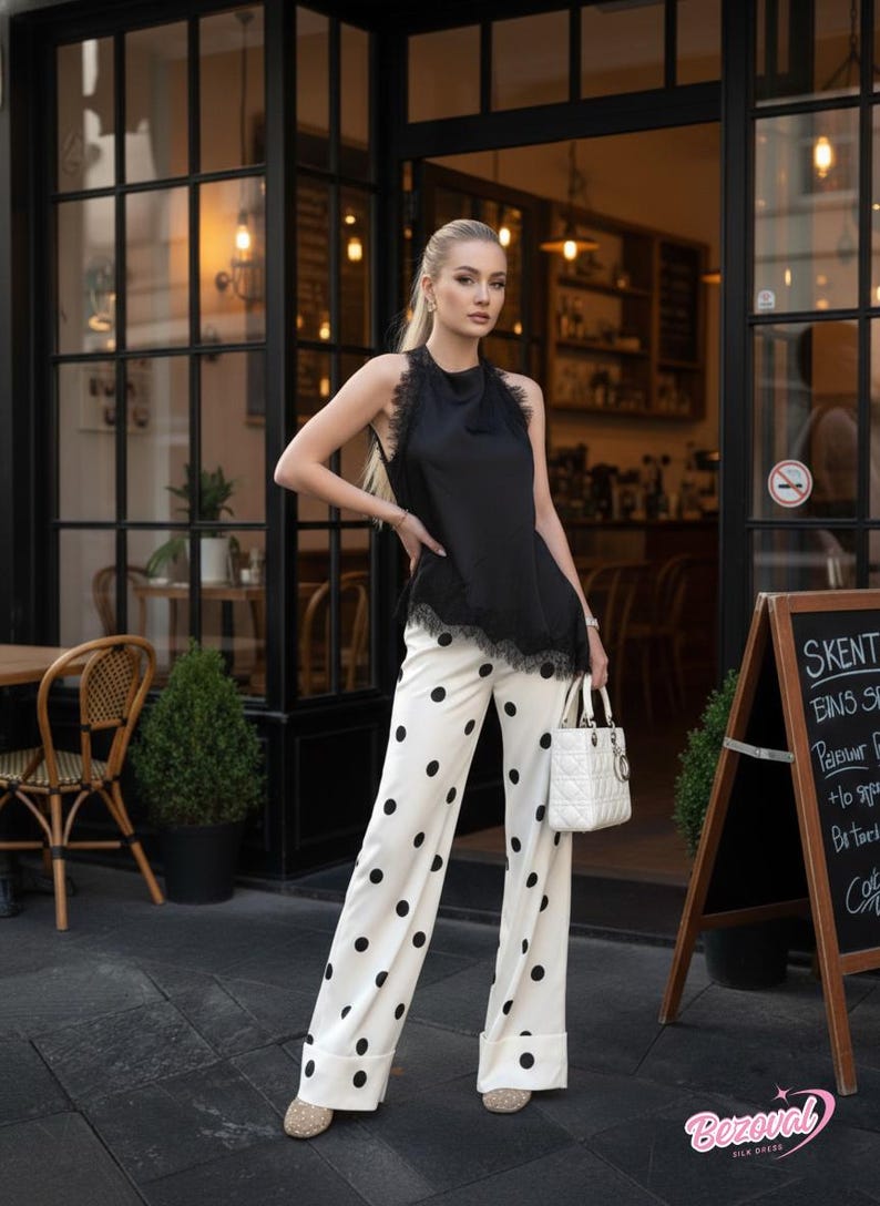 May include: A woman's outfit featuring a black sleeveless top with lace trim and white polka dot pants. The pants have a wide leg design. A white handbag and beige shoes complete the look. The background includes a cafe setting.