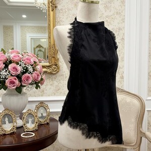 May include: A black halter top with lace trim, displayed on a mannequin. The top has a floral pattern and is set against a backdrop of a gold-framed mirror, pink roses, and antique decor.