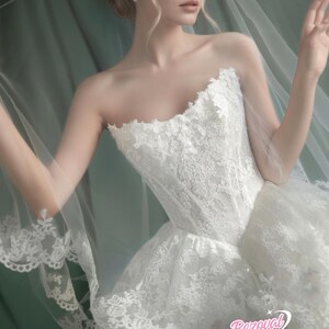 Puede incluir: Vestido de novia blanco sin tirantes con encaje y falda amplia, con velo transparente. El vestido presenta intrincados detalles de encaje floral y un corpi&ntilde;o ajustado. El velo tiene un delicado borde de encaje.