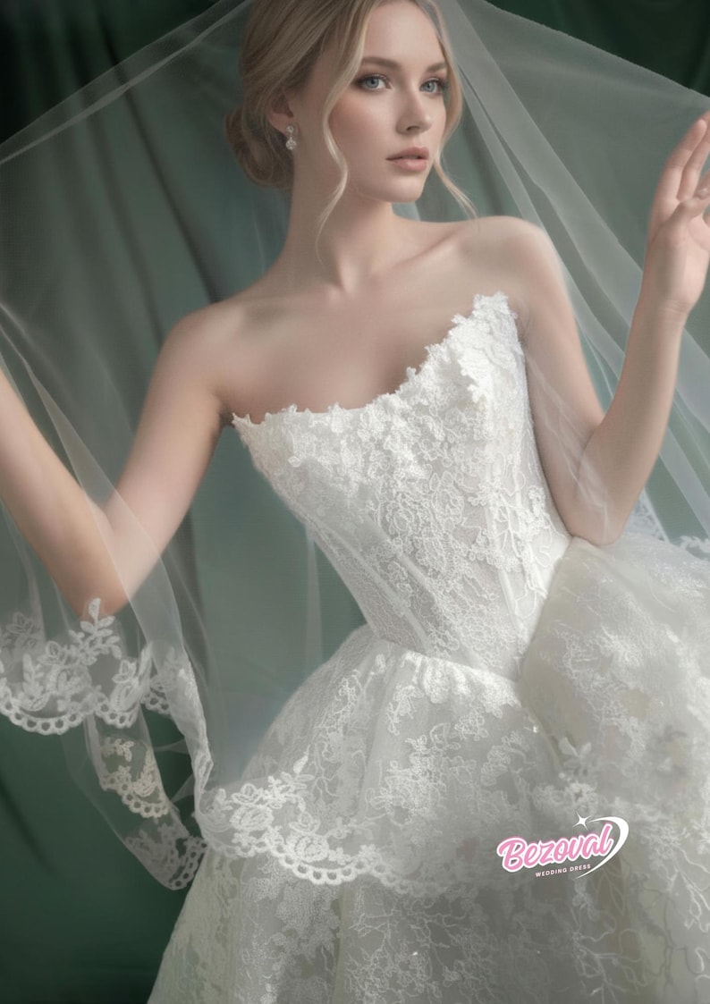 Puede incluir: Vestido de novia blanco sin tirantes con encaje y falda amplia, con velo transparente. El vestido presenta intrincados detalles de encaje floral y un corpi&ntilde;o ajustado. El velo tiene un delicado borde de encaje.