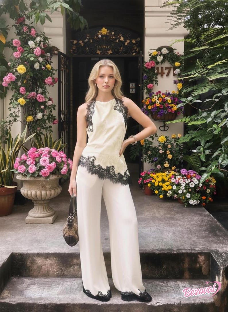 May include: Cream-colored halter top and wide-leg pants with black lace trim. The outfit is worn with black shoes and a patterned handbag. The model is standing in front of a building with floral decorations.