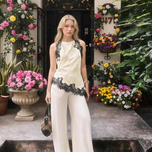 May include: Cream-colored halter top and wide-leg pants with black lace trim. The outfit is worn by a person standing on steps in front of a building with floral decorations and a brown handbag.