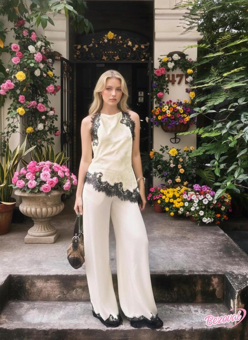 May include: Cream-colored halter top and wide-leg pants with black lace trim. The outfit is worn by a person standing on steps in front of a building with floral decorations and a brown handbag.