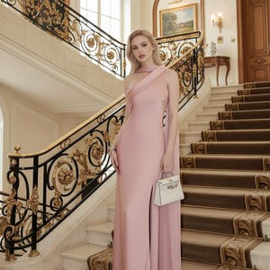 Puede incluir: Una mujer con un vestido de noche rosa p&aacute;lido de un solo hombro con una abertura alta, sosteniendo un bolso blanco. El escenario es una gran escalera con barandillas doradas y negras ornamentadas y una alfombra estampada.