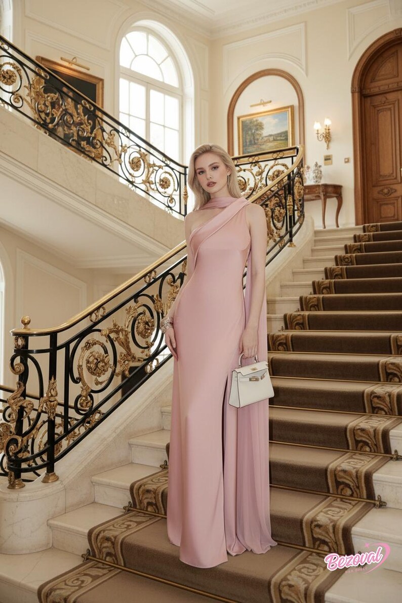 Puede incluir: Una mujer con un vestido de noche rosa p&aacute;lido de un solo hombro con una abertura alta, sosteniendo un bolso blanco. El escenario es una gran escalera con barandillas doradas y negras ornamentadas y una alfombra estampada.