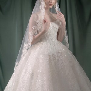 Puede incluir: Vestido de novia blanco con falda amplia y corpi&ntilde;o detallado. El vestido est&aacute; adornado con encaje y lentejuelas. Un velo transparente con ribete de encaje cae sobre los hombros. Logo "Bezoval Wedding Dress".